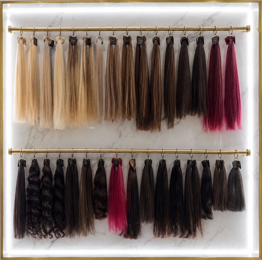 Pared iluminada de Ross Hair Studio con 32 mechones de extensiones colgados en dos barras doradas, de rubios a negros, más borgoña y fucsia